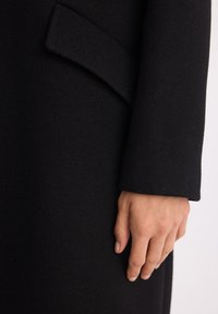Zbliżenie ręki odpoczywającej obok rękawa czarnego teksturowanego płaszcza z złożonym detalem klapki.