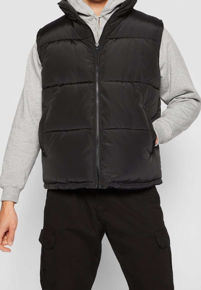 Veste matelassée noire sans manches avec fermeture éclair à l'avant, col montant et motif matelassé horizontal. Portée par-dessus un sweat à capuche gris et associée à un pantalon noir.