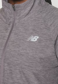 Graue sportliche Zip-Jacke mit strukturiertem Finish, ausgestattet mit einem hohen Kragen und einem weißen New Balance-Logo auf der linken Seite.