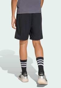 Osoba u crnim šortsevima, crnim čarapama s bijelim prugama i srebrnim Adidas tenisicama stoji, prikazana odostraga na jednostavnoj pozadini.