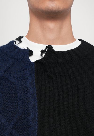 Knit sweater featuring a split design with blue cable pattern on left and solid black on right. Torn neckline with loose threads.