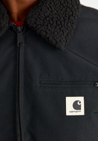 Zwarte jas met een textuurrijke zwarte fleece kraag, een rits over de volle lengte en een klein Carhartt-logo op het borstzakje.