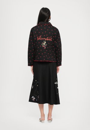 Casaco preto acolchoado com padrão de estrelas vermelhas e detalhes bordados. Apresenta um acabamento em vermelho e um colarinho oversized, usado sobre uma saia preta com bordados.