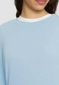 Hellblauer Pullover mit strukturiertem, Wabenmuster, rundem Halsausschnitt und sichtbarem weißen Innenkragen im Halsbereich.