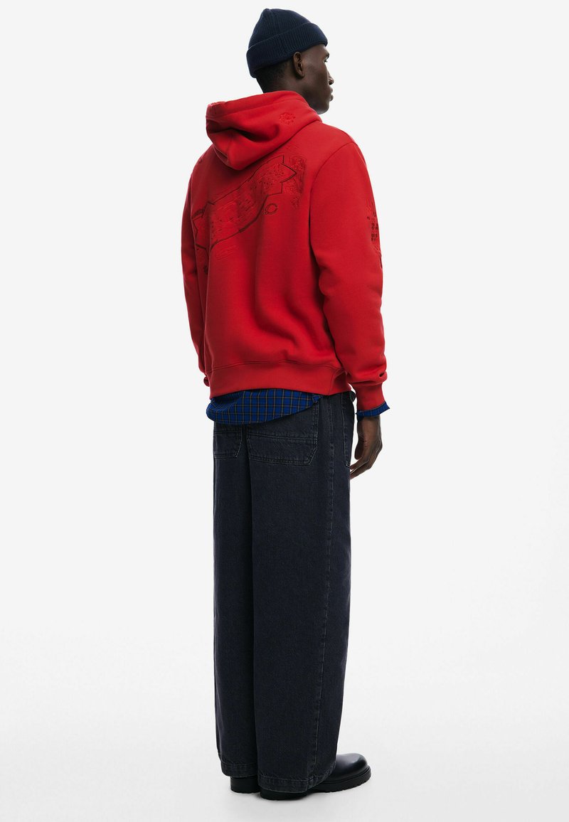 Červená mikina s kapucňou a grafickým dizajnom na chrbte, skombinovaná s modrou kockovanou košeľou a tmavými širokými džínsami. Čierna obuv.