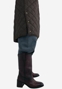 Ruskea tikattu takki nappeilla, jonka päällä on siniset farkut ja tumman burgundinpunaiset cowboy-kengät, joissa on kukkakuviot ja paksu korko.