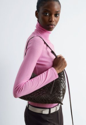 Geflochtene braune Lederhandtasche mit geschwungener Form und langem Tragegurt, gehalten von einer Person in einem eng sitzenden rosa Rollkragenpullover und dunklen Hosen.