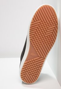 Svart sneakersula med texturerad orange gummi, med en vit kant. Förmärkt mönsterdesign ger grepp och dragkraft. Lacoste-logotyp synlig.