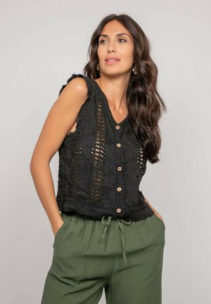 Femme aux longs cheveux ondulés portant un haut noir sans manches en crochet boutonné et un pantalon vert à cordon, les mains dans les poches.