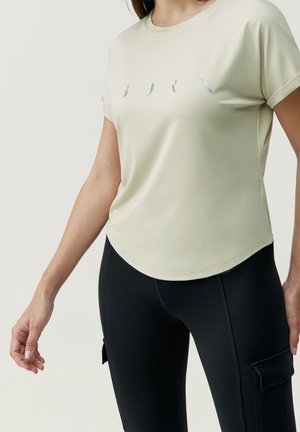 Frau trägt ein hellbeiges Kurzarmshirt mit dezentem Schriftzug und schwarze Leggings mit seitlichen Cargotaschen, steht vor einem schlichten Hintergrund.