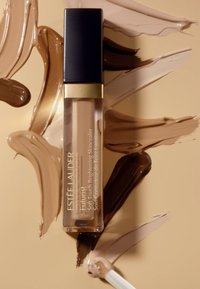 Estée Lauder Futurist Soft Touch Brightening Skincealer i en klar tube med svart kork, omgitt av swatches av foundation i forskjellige brune nyanser.