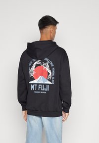 Hoodie noir avec une poche frontale, présentant un graphisme du Mont Fuji et des carpes koi au dos, souligné par des accents bleus et rouges.