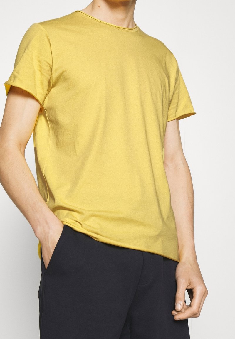 Homme portant un t-shirt jaune uni à manches courtes avec un pantalon noir, une main dans la poche et l'autre détendue sur le côté.