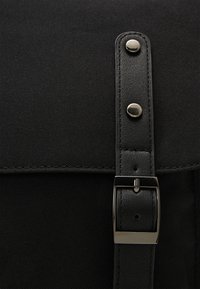 Sac à dos noir en tissu texturé avec une sangle en cuir brillant. Il comporte des accessoires argentés et un détail de boucle rectangulaire.