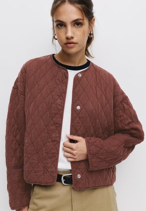 Jachetă matlasată de culoare cărămizie, cu guler rotund, nasturi tip snap și umeri lăsați, asortată cu un tricou alb și pantaloni bej.