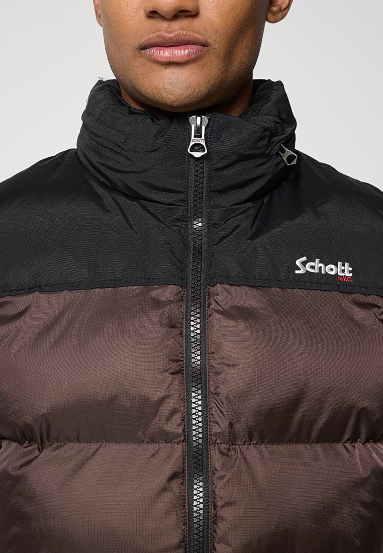 Veste rembourrée noire et brune avec col montant, fermeture éclair sur toute la longueur à l'avant et logo sur le côté gauche. Présente un tissu texturé avec des coutures horizontales.