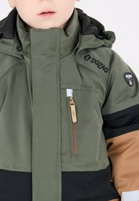 Grøn jakke med sorte og brune accenter, lynlås foran, høj krave og logoer på brystet. Tekstureret stof med et sporty design.