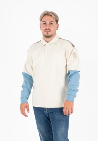Crème polo shirt met blauwe lange mouwen, voorzien van een borstzak en een schouderaccent in bruin. Inclusief een ronde logo patch. Jeans zichtbaar.