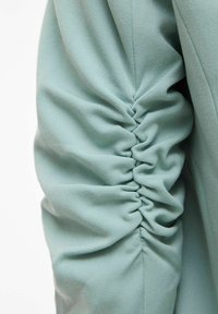 Manchon en tissu vert clair avec des détails froncés le long de la couture du milieu, créant des plis et des creux texturés.