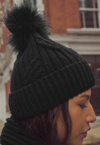 Schwarze gestrickte Mütze mit einem Rippmuster, ausgestattet mit einem schwarzen Kunstfell-Pompon oben. Die Textur ist dick und gemütlich, geeignet für kaltes Wetter.