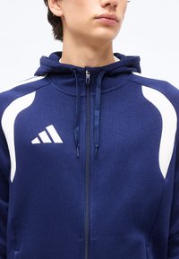 Νεαρό άτομο που φοράει μπλε σκούρο φούτερ με φερμουάρ, λευκές λεπτομέρειες και αφηρημένο λευκό λογότυπο στο αριστερό στήθος.