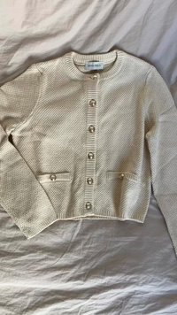 Világos bézs színű kardigán, strukturált kötött anyagból készült, kerek nyakkal, nyolc díszgombbal és két elülső zsebbel.