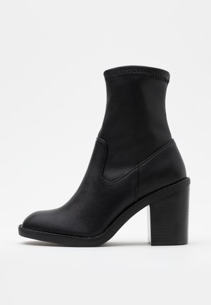 Bottine en cuir noir avec talon haut, texture lisse, bout arrondi et tige sans coutures. Présente un accent de col cousu.