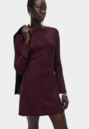 Kurzes burgunderrotes Kleid mit glitzernder Textur, langen Ärmeln und weitem Rundhalsausschnitt. Das Model hält einen schwarzen Schal, der ein minimalisches Design aufweist.