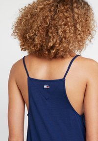 Granatowy top na ramiączkach z cienkimi paskami, z trójkątnym wzorem na plecach oraz małą metką z logo przy dekolcie. Miękka, teksturowana tkanina.