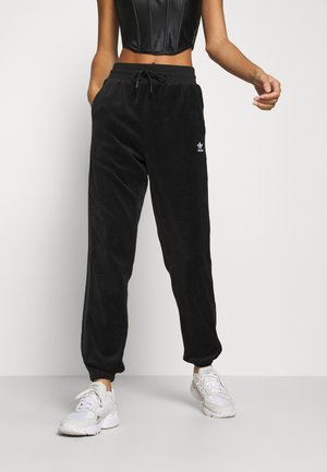 Femme portant un jogging Adidas noir, des baskets blanches et un top court noir sur un fond clair uni.