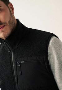 Czarna polarowa kamizelka z podwyższonym kołnierzem, teksturowany materiał, pełny zamek błyskawiczny oraz kieszeń na piersi na zamek. Noszona na szarym t-shircie.
