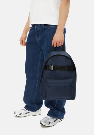 Osoba oblečená v volných modrých džínách a bílých teniskách, držící tmavě modrý batoh s přední kapsou a přezkou za rukojeť.