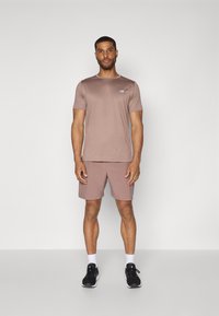 Kortermet, lysebrun treningstrøye med en liten logo, kombinert med matchende shorts. Sorte sneakers og hvite sokker fullfører antrekket.
