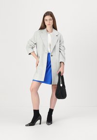 Blazer oversize grigio chiaro, blouse bianca, mini gonna blu, calze nere alla caviglia e borsa a tracolla nera intrecciata. Scarpe nere con tacco e dettagli lucidi.