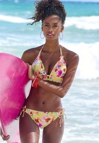 Bloem bikini met een gele achtergrond, voorzien van roze accenten en strikken aan de zijkanten. Roze surfboard onder de arm, tegen een oceaan achtergrond.