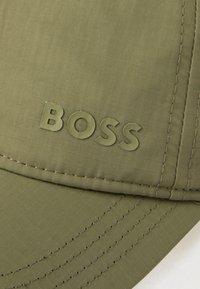 Kapuca olivno zelene barve iz teksturirane tkanine, z izbočenim logotipom "BOSS" v enaki barvi ter vidnimi šivi na robu.