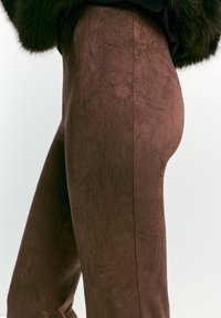 Primo piano di una persona che indossa pantaloni brown a vita alta con una texture e una giacca scura con rifiniture in pelliccia su uno sfondo semplice chiaro.