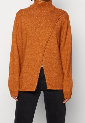 Pull en maille orange rouille à col montant avec manches longues et fente diagonale à l'avant, porté avec un pantalon noir taille haute.