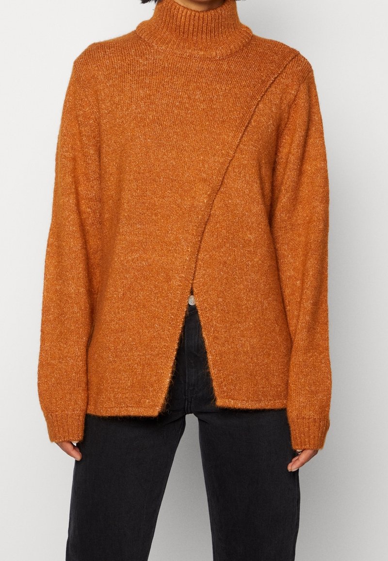 Pull en maille orange rouille à col montant avec manches longues et fente diagonale à l'avant, porté avec un pantalon noir taille haute.