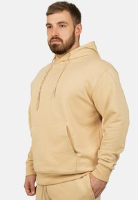 Felpa beige realizzata in tessuto morbido, con tasca frontale, coulisse e polsini a coste. Il design è semplice, senza motivi visibili.