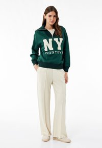Sudadera verde con el texto blanco "NY DOWNTOWN", cuello de cremallera y puños acanalados, combinada con pantalones de pierna ancha color crema y zapatos deslizables beige.