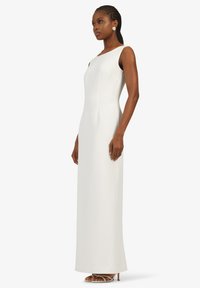 Robe longue blanche au design épuré et ajusté, texture lisse, larges bretelles et une subtile fente sur le côté. Associée à des sandales blanches à lanières.