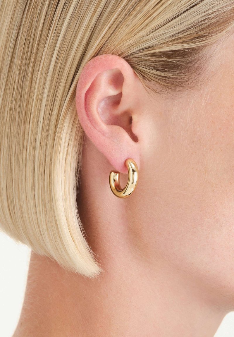 Personne blonde aux cheveux courts portant une boucle d'oreille anneau épais et brillant en or à l'oreille gauche, vue en gros plan.