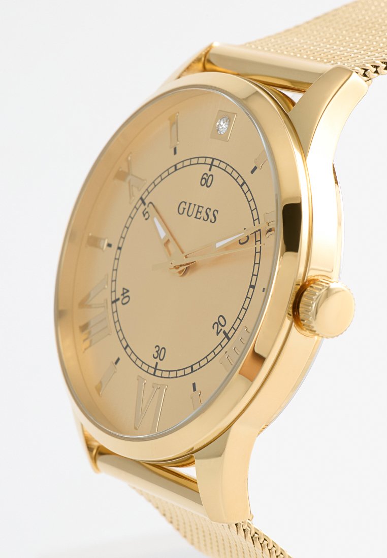 Montre bracelet Guess en or avec un bracelet en maille, chiffres romains, détail en cristal à 12 heures et marquages des minutes à l'intérieur du cadran.