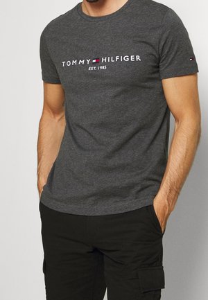 Homme portant un t-shirt gris foncé Tommy Hilfiger avec logo et pantalon noir, mains dans les poches, sur un fond clair uni.