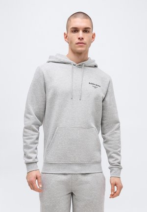 Mladý muž s krátkymi vlasmi v svetlosivej mikine Björn Borg a zodpovedajúcich teplákoch na bielom pozadí.