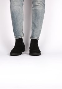 Bottines en suédine noire avec un bout arrondi et une semelle en caoutchouc, associées à un jean en denim bleu clair retroussé. Design simple.