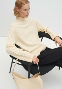 Beiger Strickpullover mit hohem, geripptem Kragen, weiten Ärmeln und geripptem Saum; kombiniert mit schwarzer Hose und einer hellcremefarbenen Handtasche.