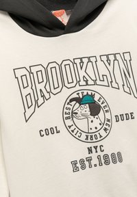 Hoodie in crème en zwart, met de tekst "Brooklyn" en een grafische afbeelding van een hond in een hoed. Gemaakt van zachte stof met een gestructureerde afwerking.