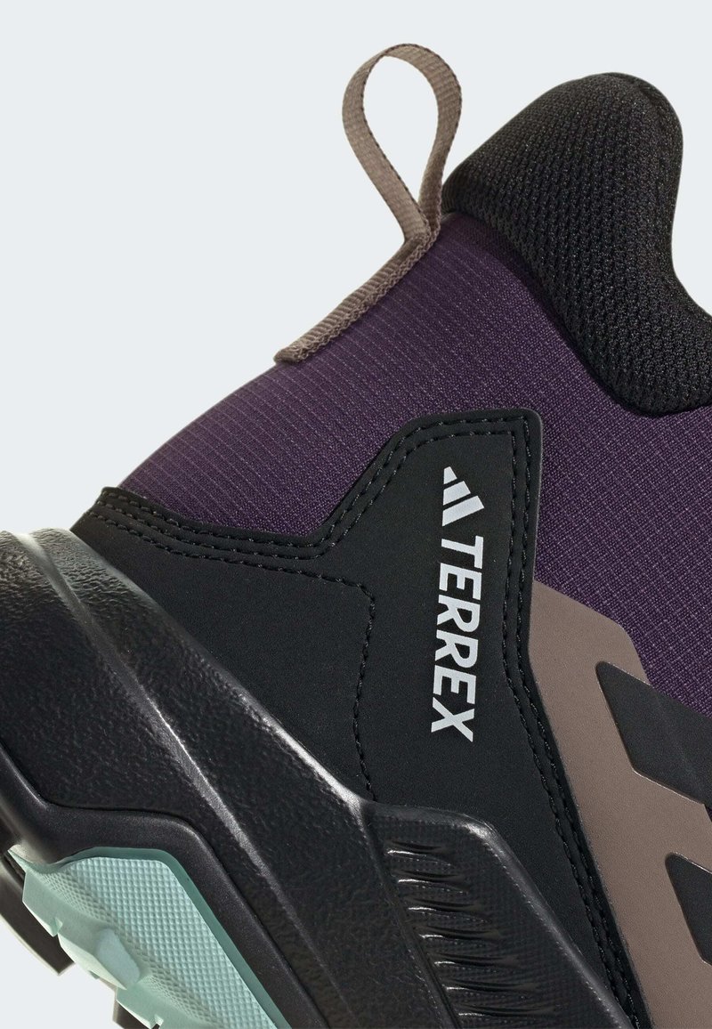 Zapato de senderismo negro y morado con parte superior texturizada, suela de goma y un destacado diseño con la marca 'TERREX'. Cuenta con un lazo de tela para ponerlo y quitarlo fácilmente.